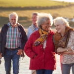 Group Of Smiling Senior Friends Walking Arm In Arm Along Shoreline Of Winter Beach